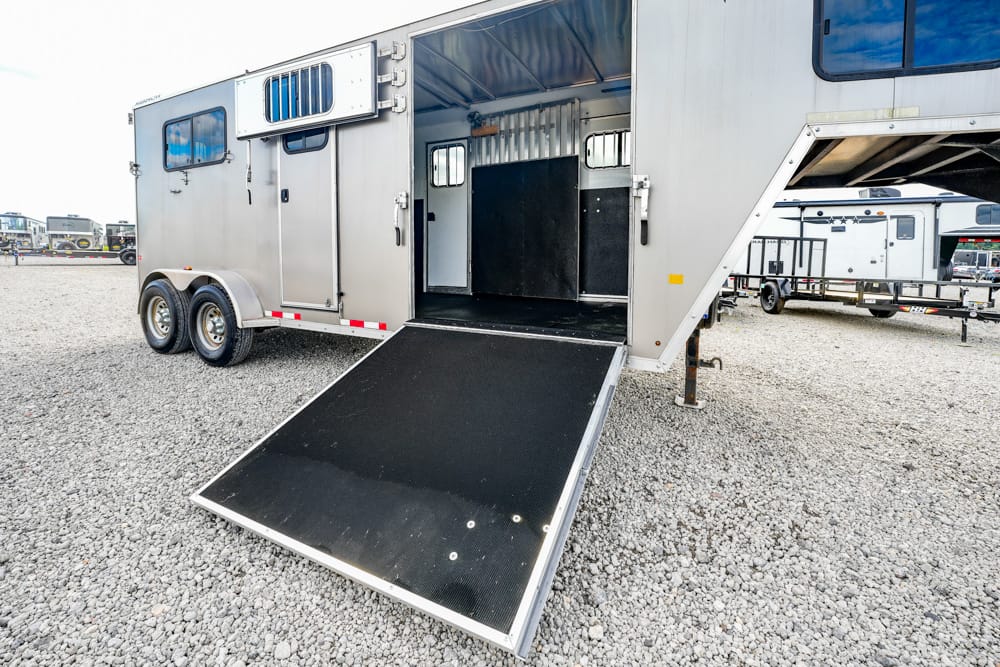 Aluminum horse trailer with its rear ramp lowered for loading, interior stalls visible on a gravel lot.