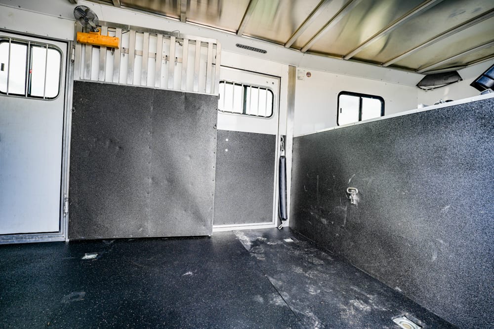 Interior of a horse trailer: narrow stall with dark divider panels, white walls, metal bars near the ceiling, and a worn black rubber floor with scuffs and a small window in the background