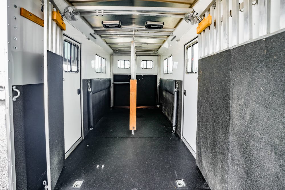 Interior of a horse trailer with a central wooden partition and padded side walls, empty stalls, and small windows along the sides to vent.