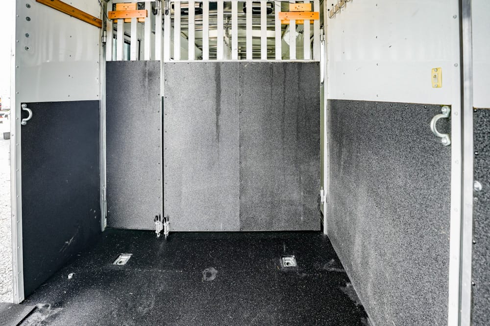 Interior of a metal horse trailer with partition panels and padded walls, showing a dark rubber floor and tie-down anchors for cargo or animals