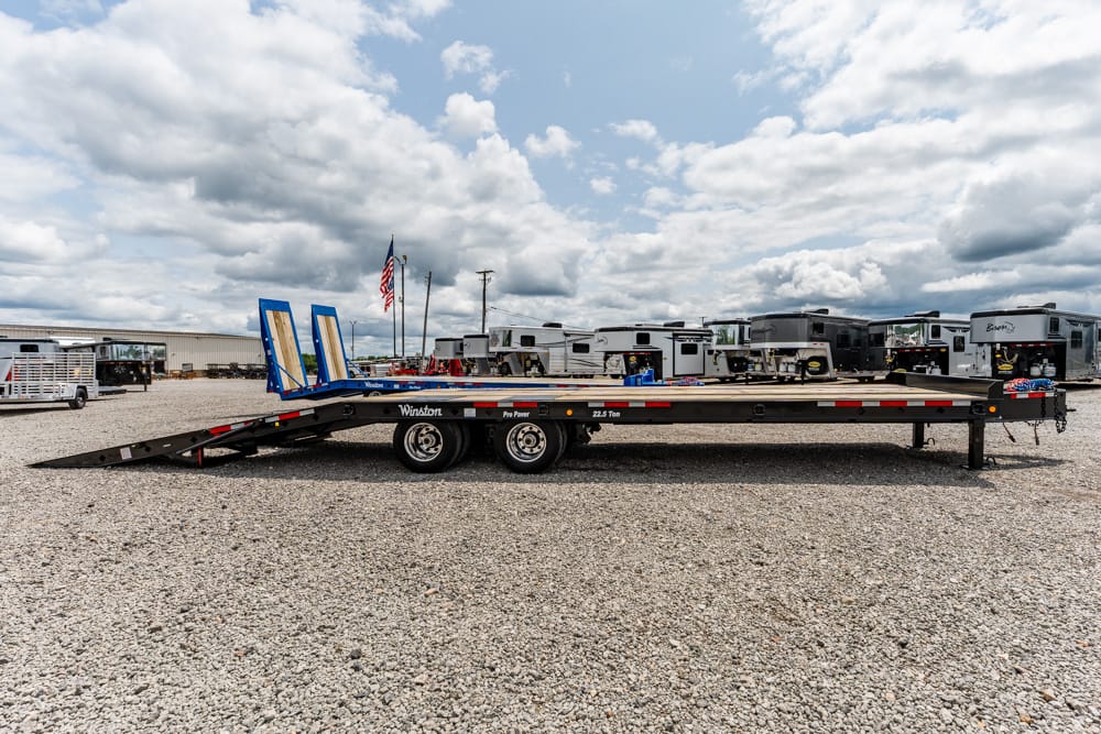 Long flatbed trailer with dual axles and rear loading ramps in a gravel lot filled with RVs and trailers under a cloudy sky