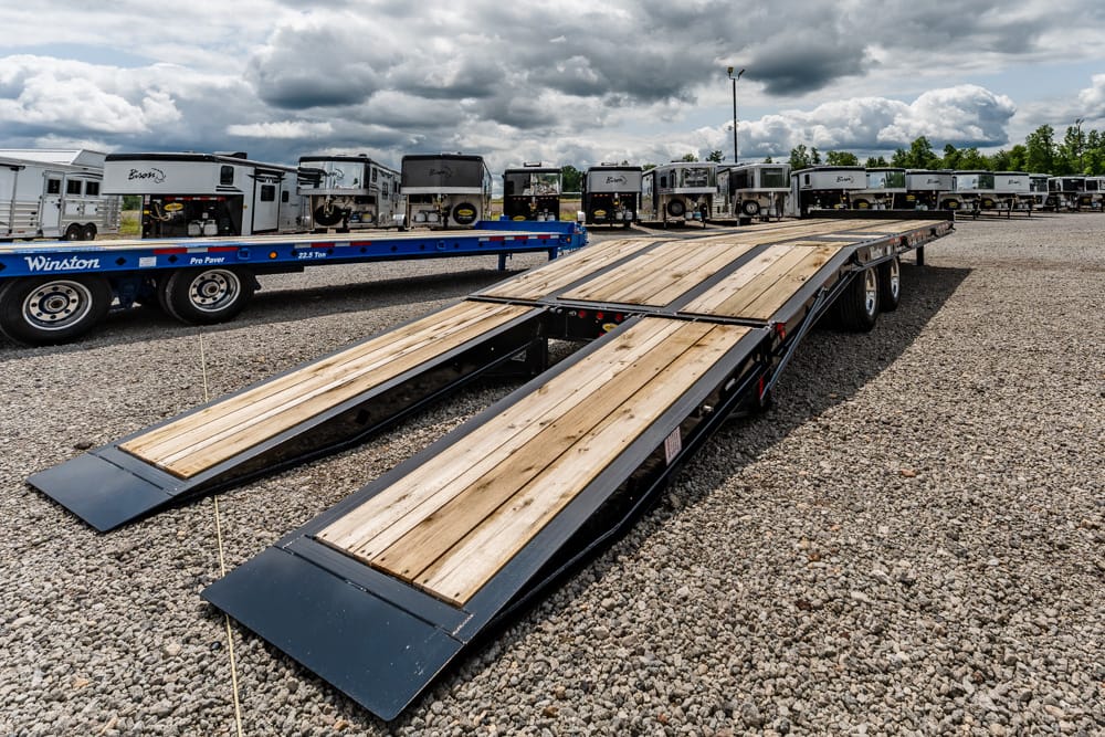 Flatbed trailer with lowered loading ramps on a gravel lot, several trucks parked in the background under a cloudy sky.
