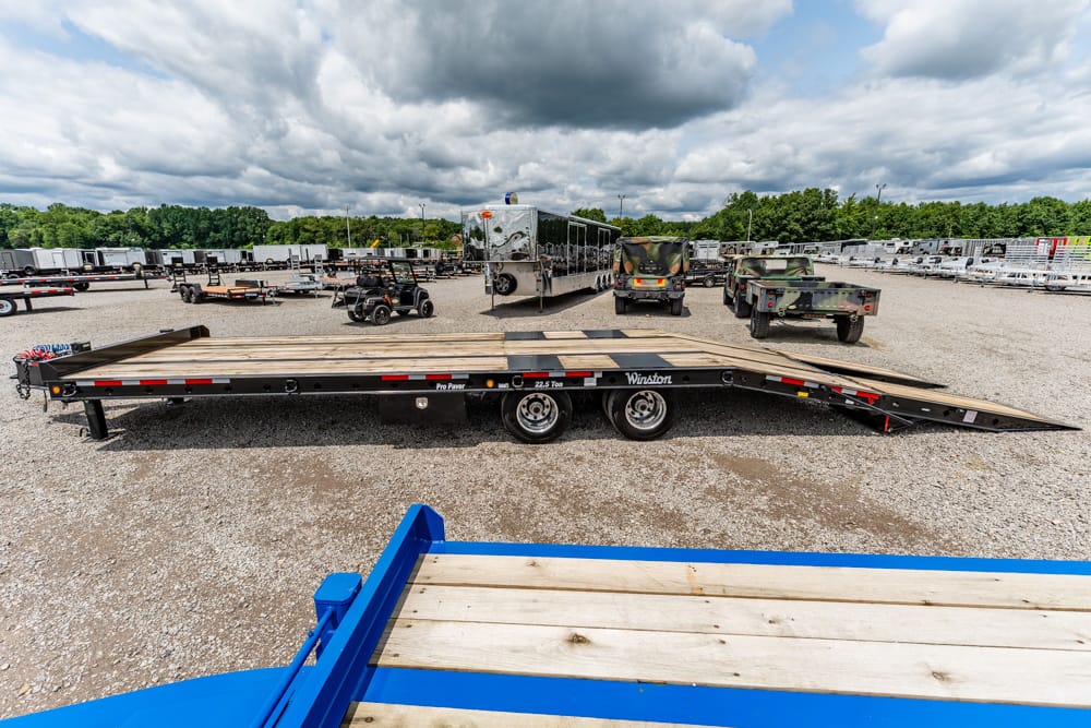 Flatbed trailer with a wooden deck in a gravel trailer yard, with additional trailers and equipment in the background under a cloudy sky and trees far away.