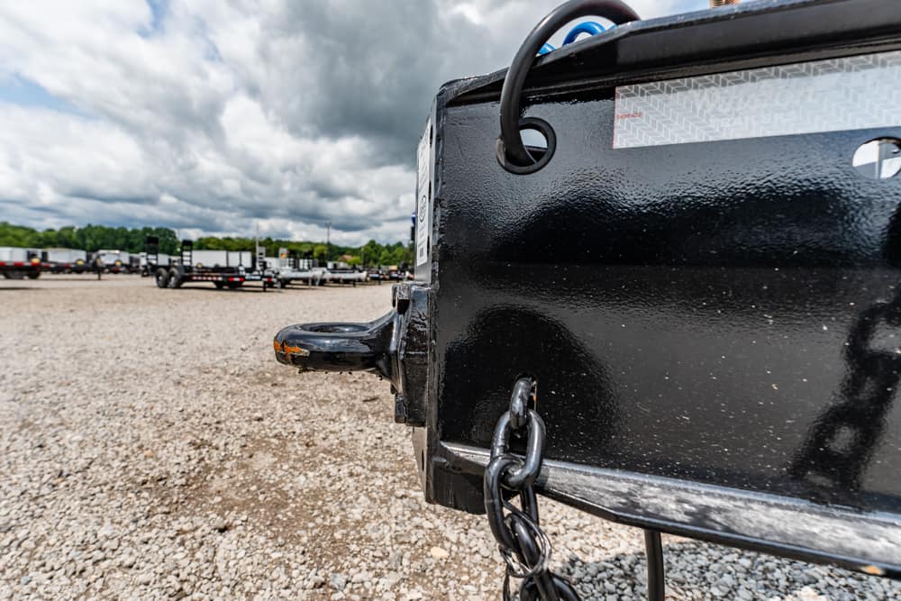Close-up of a black trailer hitch on a gravel lot with other trailers in the background and a cloudy sky above.