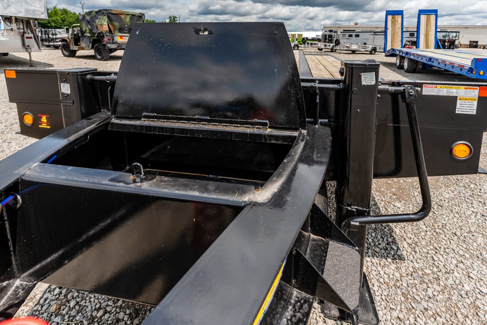 Heavy-duty black trailer with an open cargo bed and supporting beams, parked on a gravel lot with other trucks in the background.