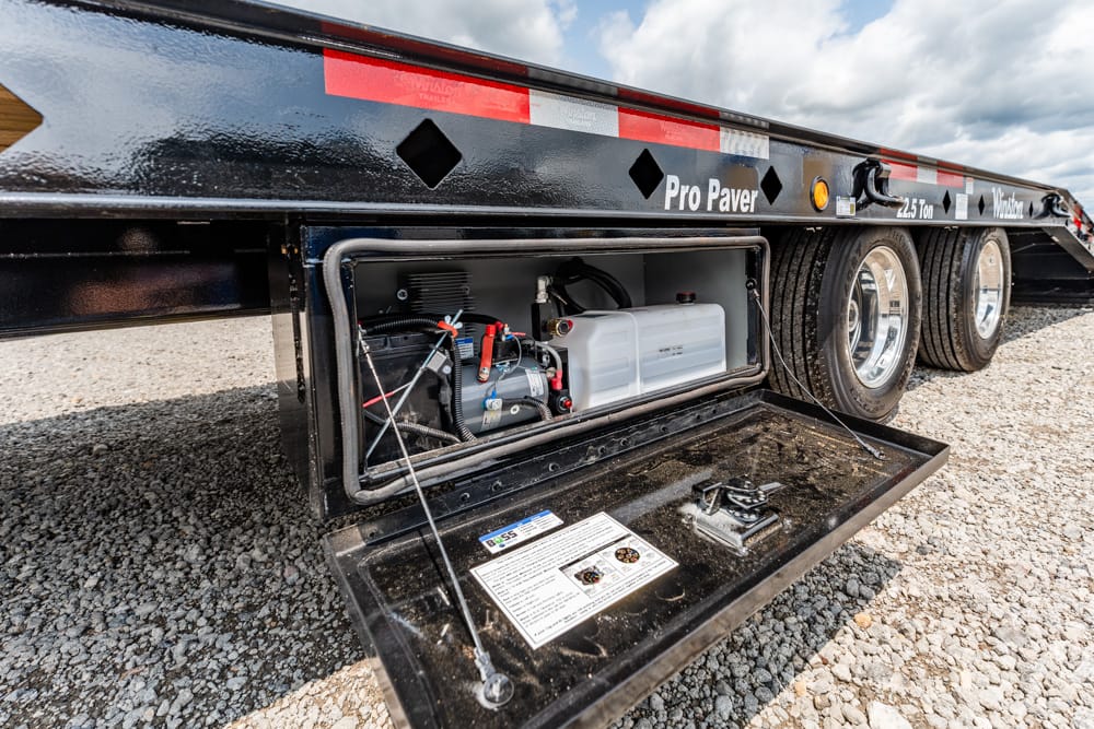 Side-view of a Pro Paver trailer with its storage hatch open, revealing a white fuel tank and small engine with hoses inside the compartment on a gravel surface.