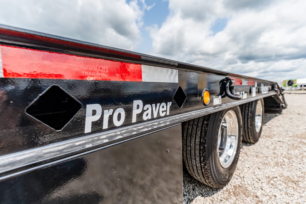 Close-up of a black Pro Paver trailer side with red reflective stripe and two axles on a gravel lot, wheels visible.