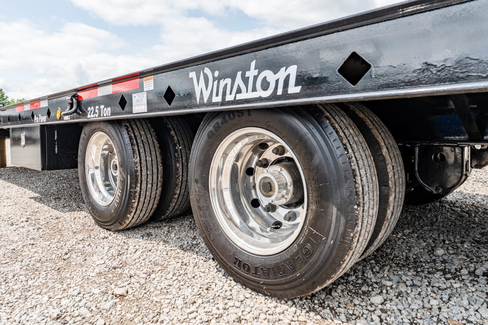 Close-up of dual wheels on a Winston flatbed trailer on a gravel lot, showing chrome rims and treaded tires.