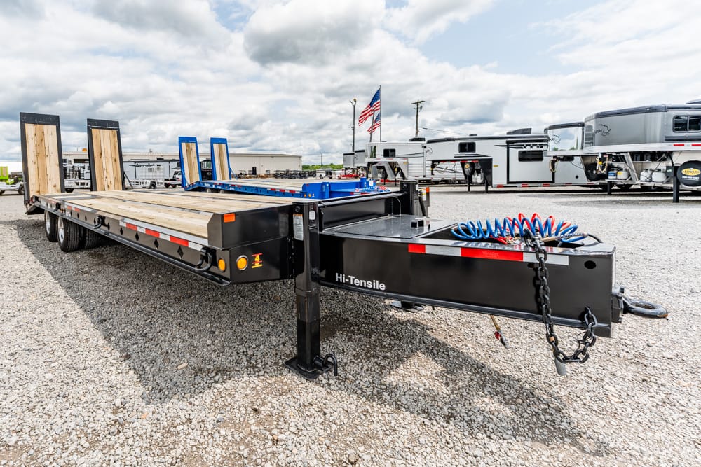 Long black flatbed trailer with a wooden deck and blue loading ramps on a gravel lot, labeled Hi-Tensile.