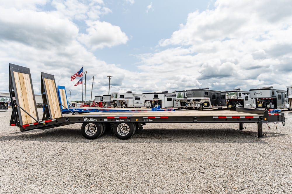 Long flatbed trailer with loaded ramp extensions in a gravel RV storage lot, American flag in the background