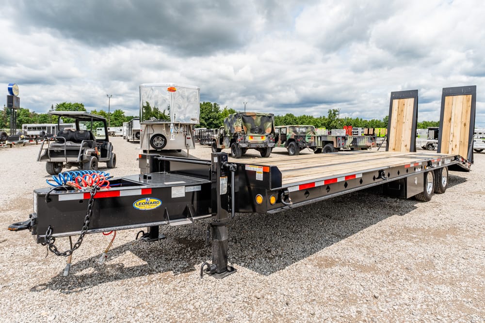 Long flatbed trailer with wooden deck and dual rear ramps parked in a gravel lot, ready for hauling equipment.