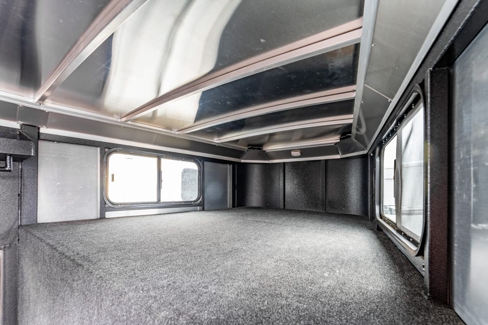 Interior of a small camper/truck bed with carpeted floor, metal walls, and side windows viewed from the back end.