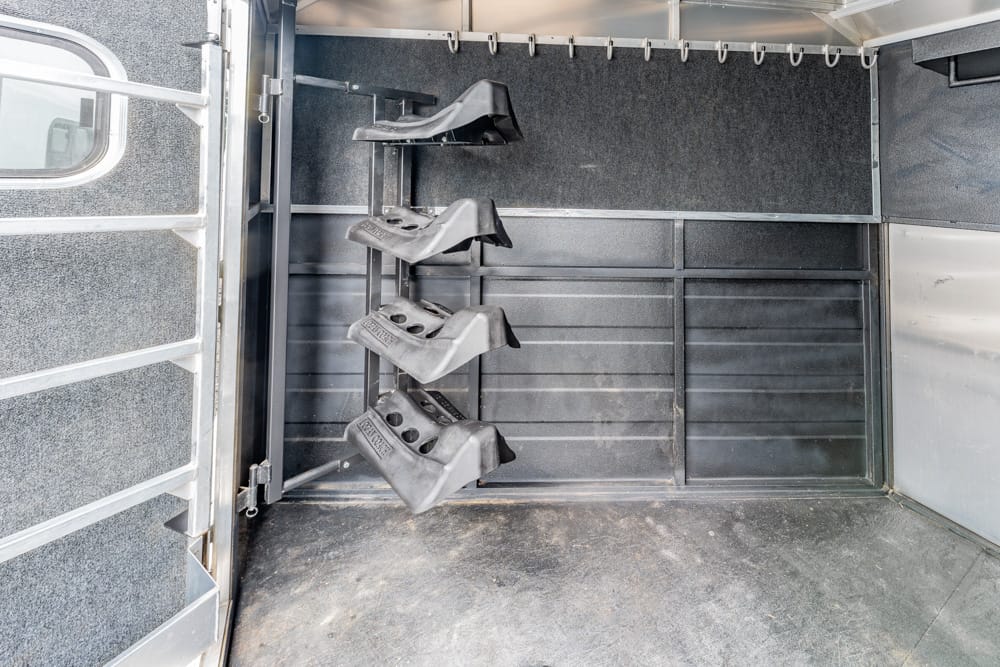 Interior of a horse trailer showing a vertical rack with four saddle racks hanging on the wall, plus a rubberized floor and side window.