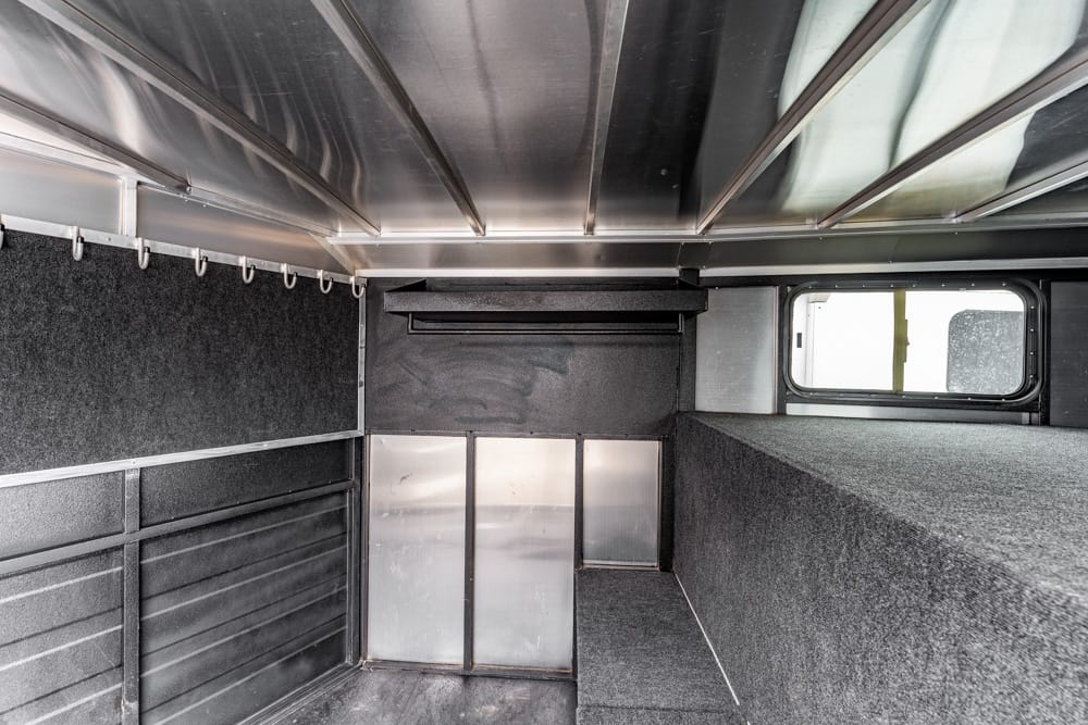 Interior of a dark cargo trailer with a raised bunk on the right, metal wall panels, and hooks along the left; a small window at the far end.
