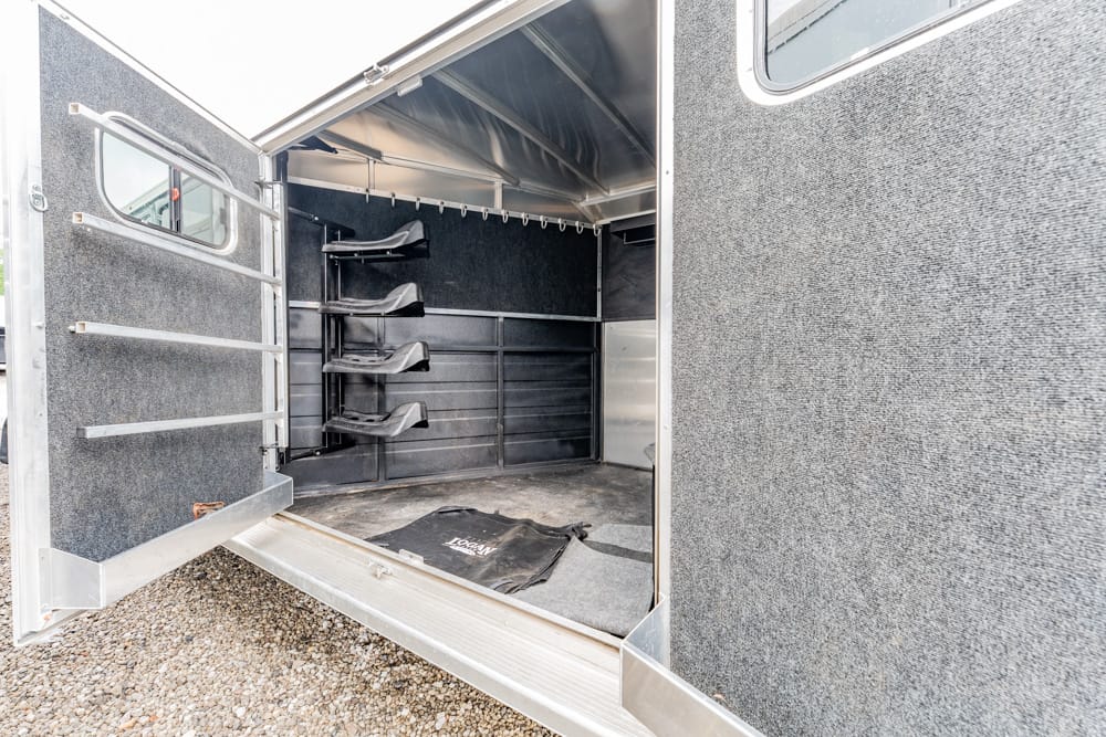 Interior of a horse trailer with four saddle racks along the left wall, gray padded interior, and an open rear door.