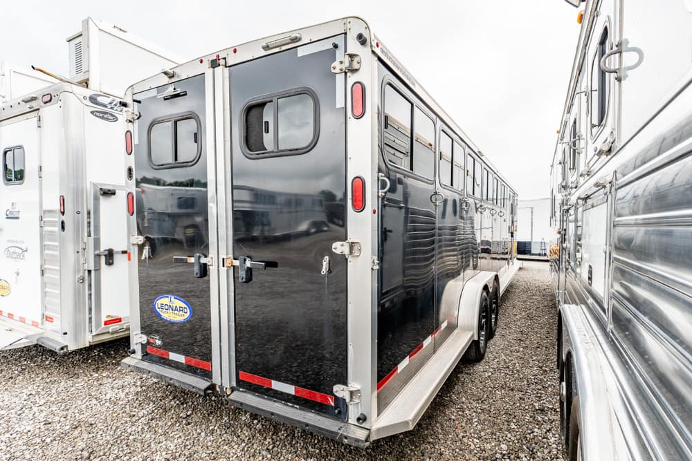 Black enclosed horse trailer with dual axles and windows parked among other trailers on a gravel lot.