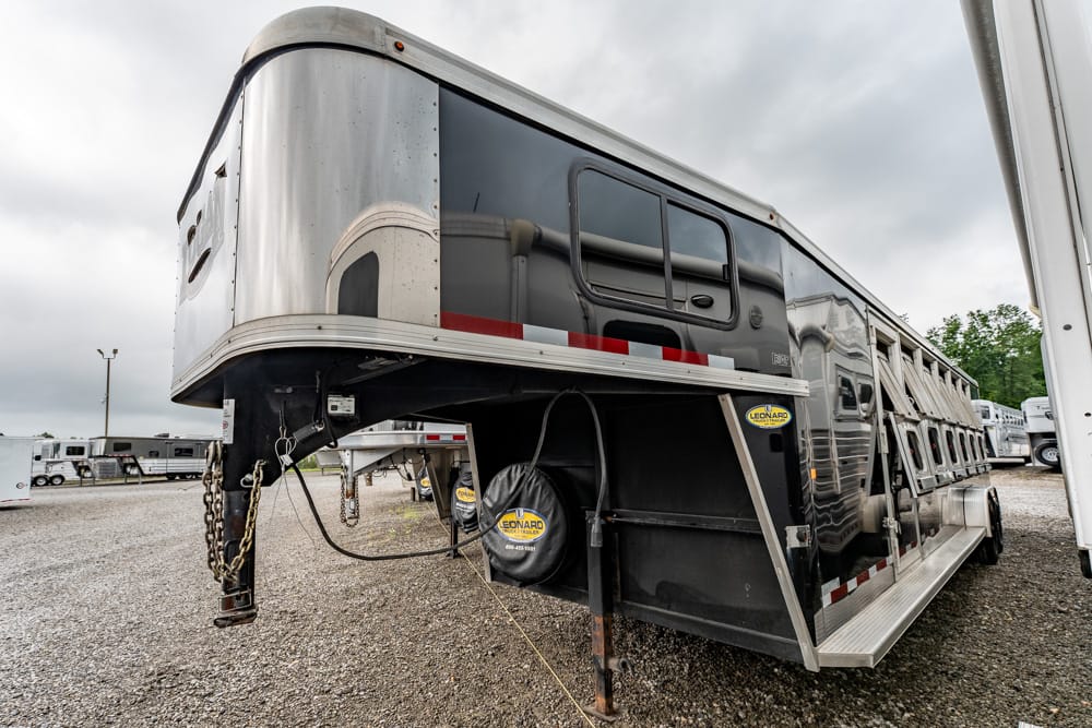 A long silver horse trailer with a black lower section and a side loading ramp, parked on gravel in a lot.