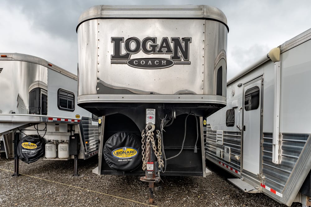 Front view of a silver Logan Coach trailer in a line of horse trailers, with hitch and chains visible on a gravel lot.