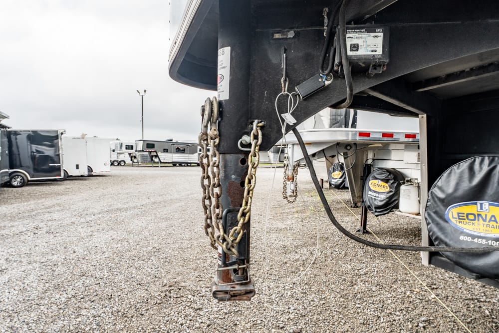 Close-up of a trailer hitch with rusty chains and electrical cables on a black truck trailer in a gravel lot.