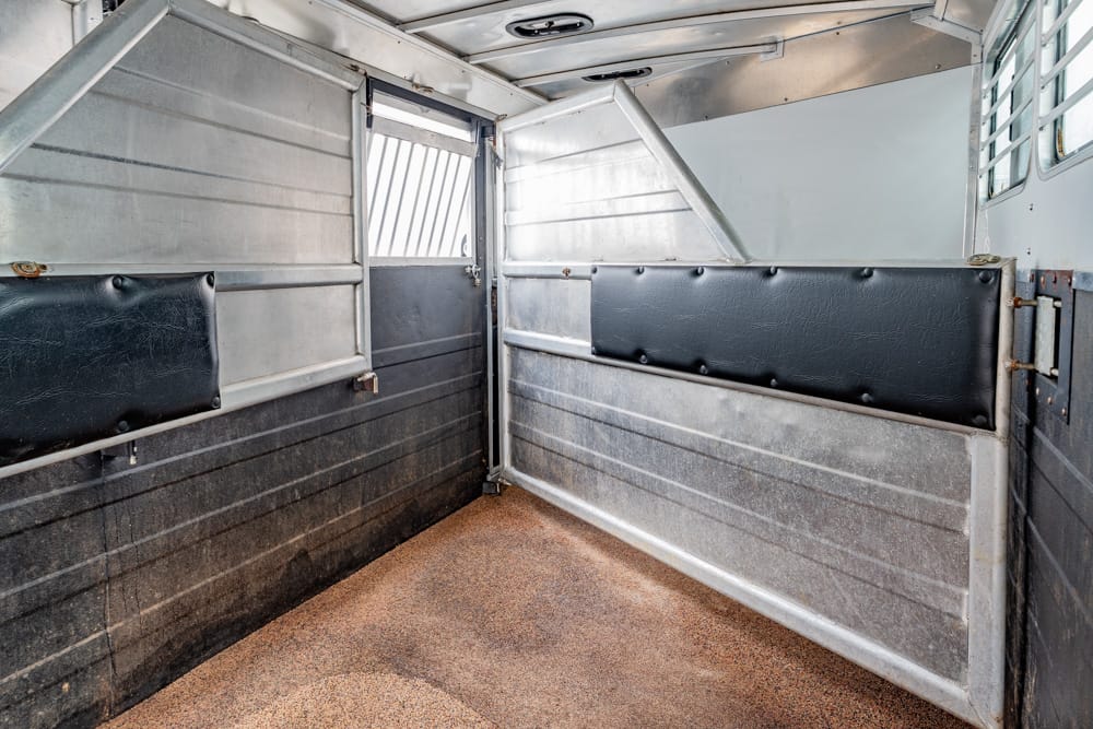 Empty horse trailer stall with metal walls and padded dividers, sawdust floor.