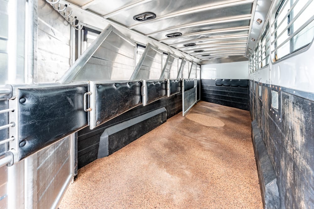 Interior of an empty horse trailer with black padded stall dividers along the left side and a tan textured floor