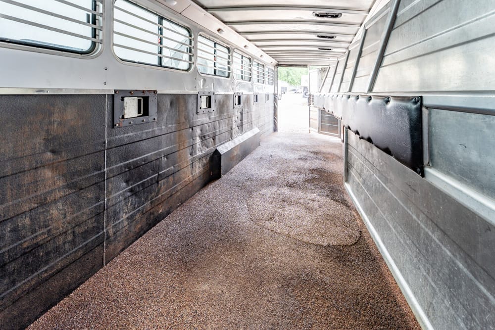 Interior of an empty, rusty metal bus or trailer with window slats on the left and padded wall on the right, long narrow aisle toward daylight at the end.