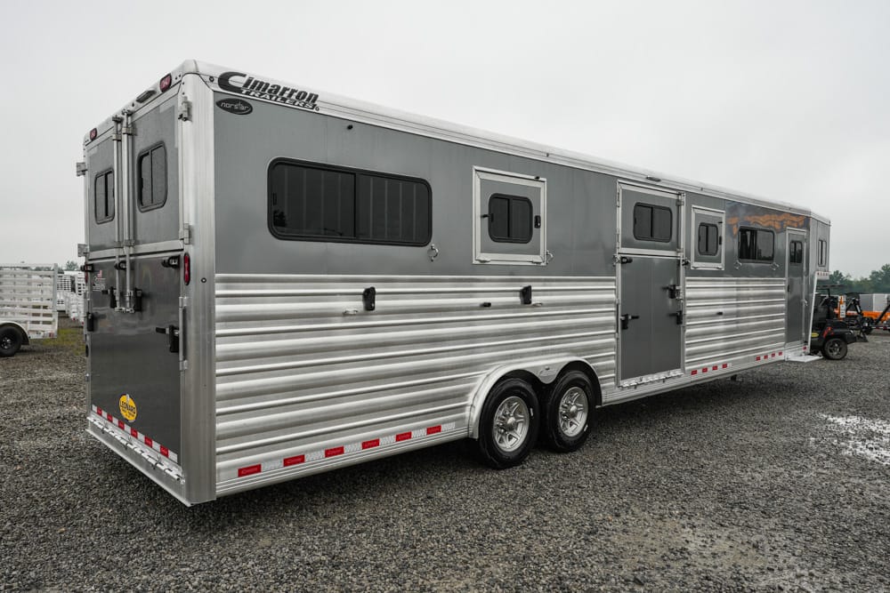 Silver aluminum horse trailer with two axles, side windows, and multiple entry doors on a gravel lot.