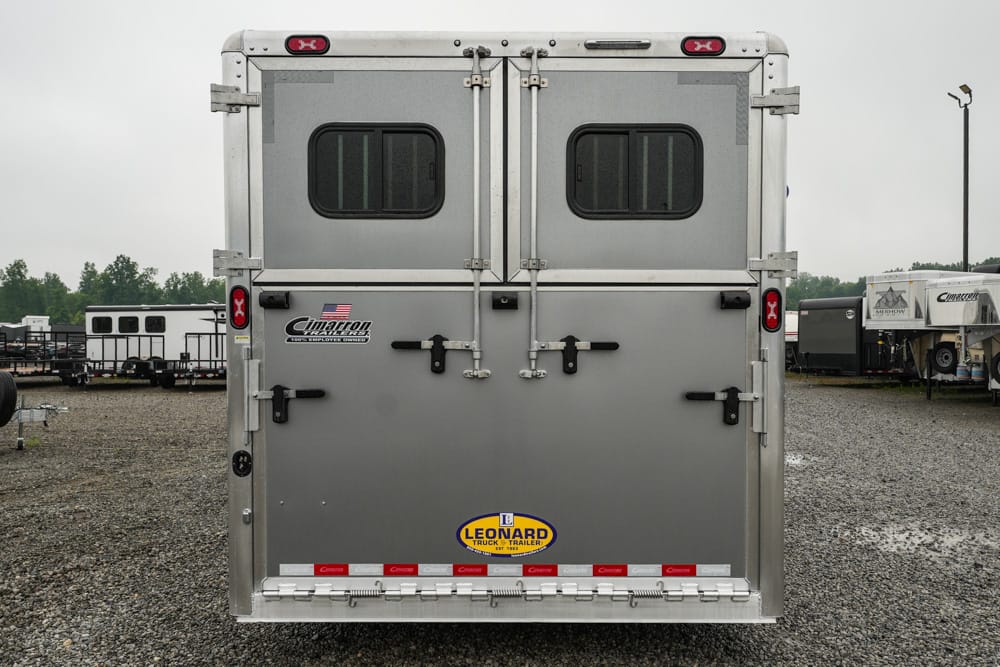 Rear view of a silver livestock trailer with double doors, latches, and small windows; brand stickers and a Leonard trailer logo visible on a gravel lot.