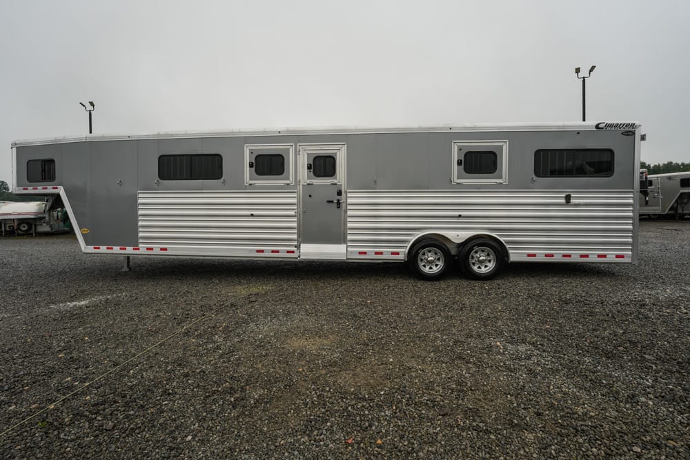Long silver horse trailer with living quarters, side windows, and dual axles parked on a gravel lot in overcast weather.