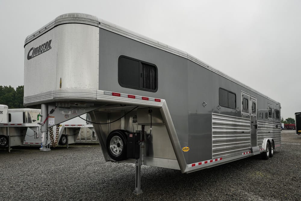 Grey and white gooseneck horse trailer with living quarters, parked on gravel. The front hitch area and spare tire are visible, with multiple side windows and a silver corrugated paneling section.