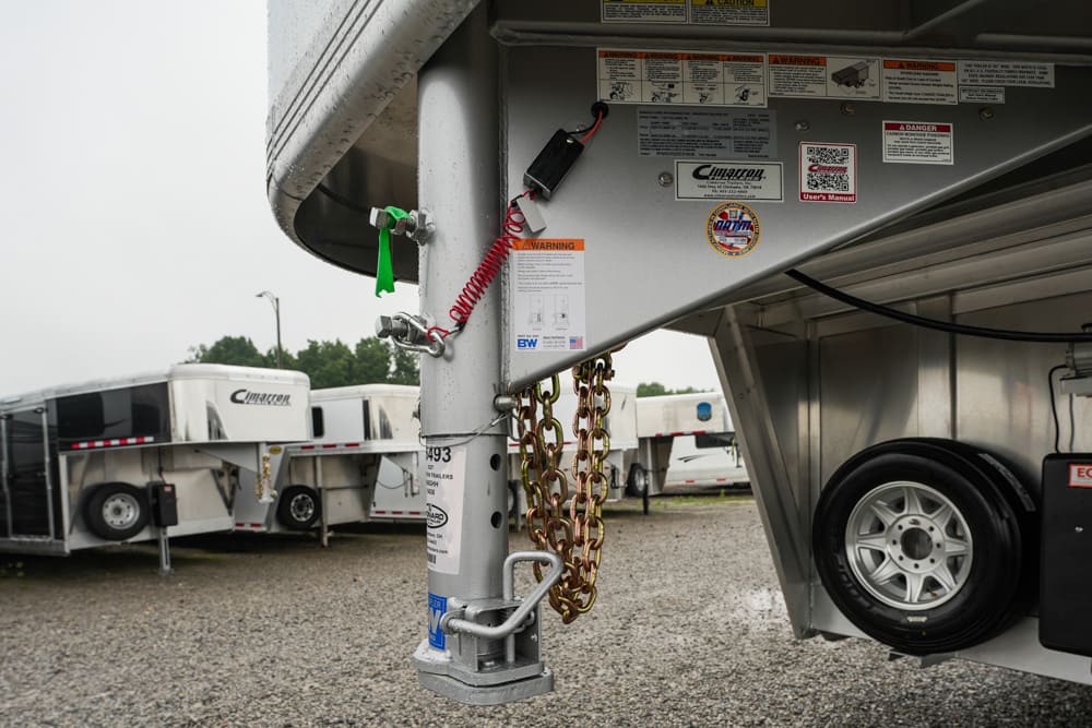 Close-up of a trailer hitch with safety chains, warning labels, and a red coiled cord; other trailers visible in the background.