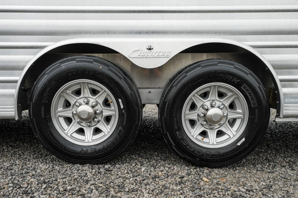Pair of silver trailer wheels with black tires mounted on a ribbed aluminum trailer body, parked on gravel.
