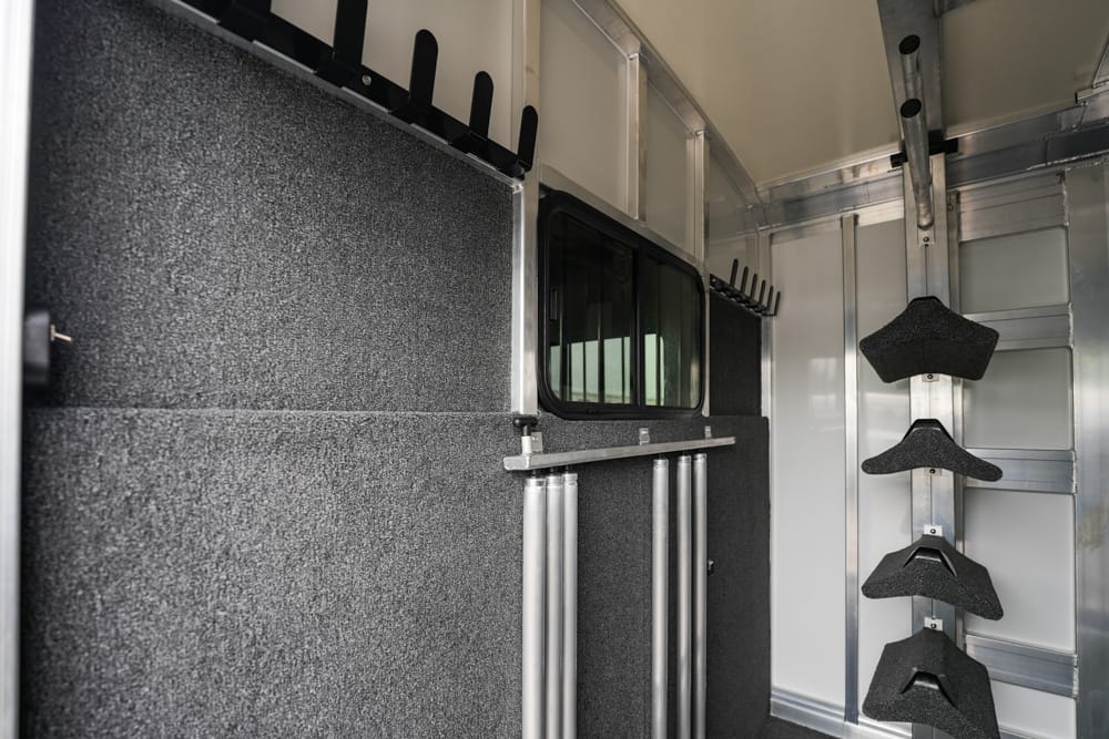 Interior of a horse trailer featuring padded partition, silver rails, and wall-mounted saddle racks along the right side, with a small window in the partition.