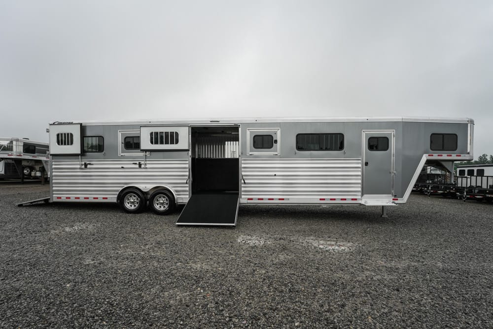 Long silver horse trailer with a lowered ramp in a gravel lot under a cloudy sky