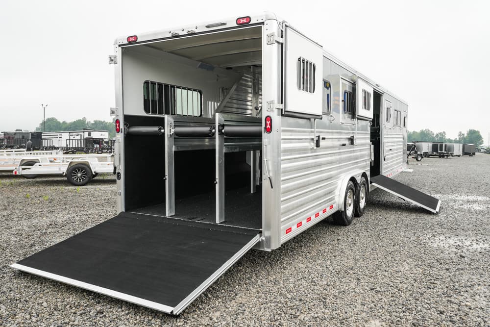 Silver horse trailer with ramp down and dividing stalls, parked on a gravel lot.