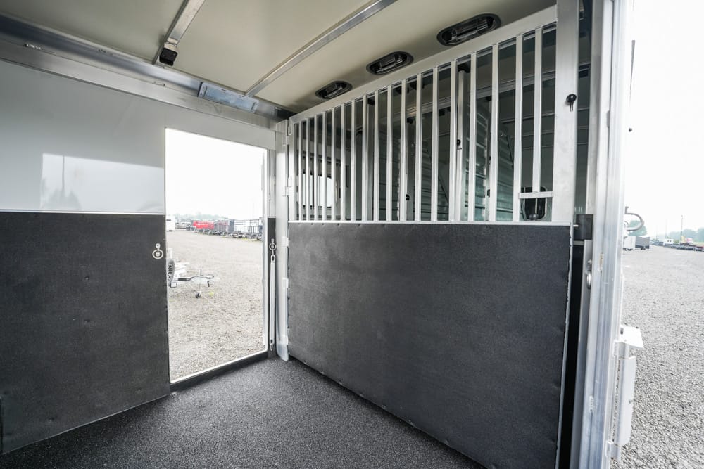 Interior of a horse trailer with divider, padded lower walls, and an open doorway to a gravel lot outside.