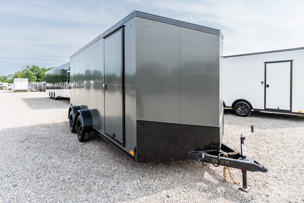 Gray enclosed cargo trailer with diamond-plate front, attached to a hitch, parked on a gravel lot.