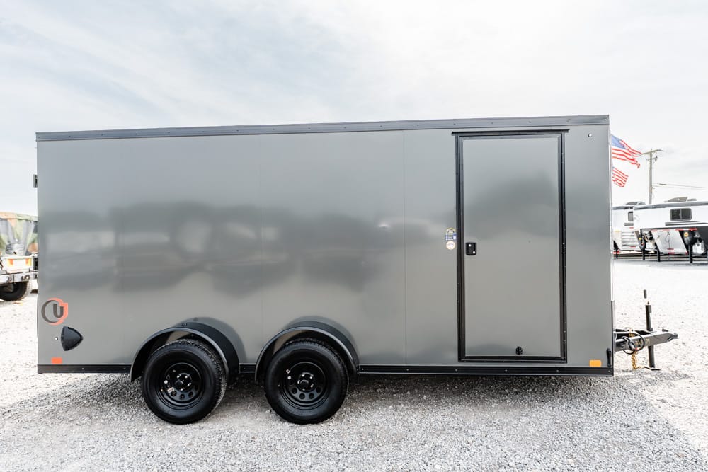 Gray enclosed cargo trailer with dual axles and a side access door on a gravel lot, hitch at front and small logo near rear wheel.