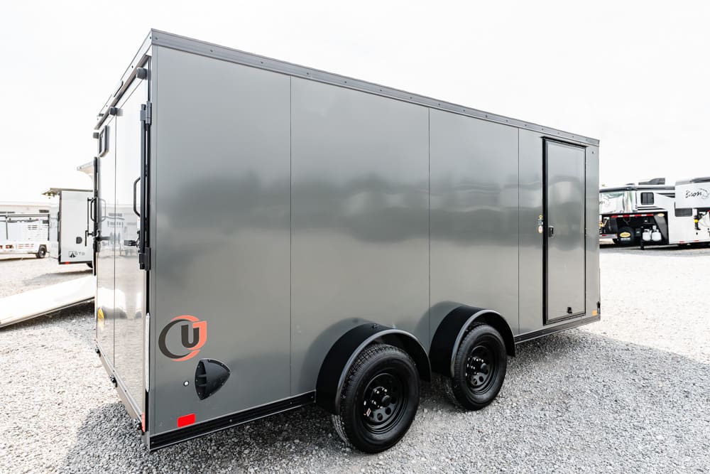 Gray, enclosed dual-axle cargo trailer on a gravel lot, side door closed and logo near the rear corner.
