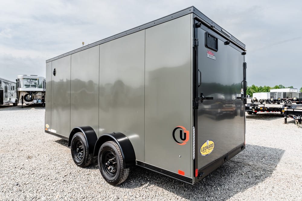 Gray enclosed cargo trailer with dual axles parked on a gravel lot; back doors shut.