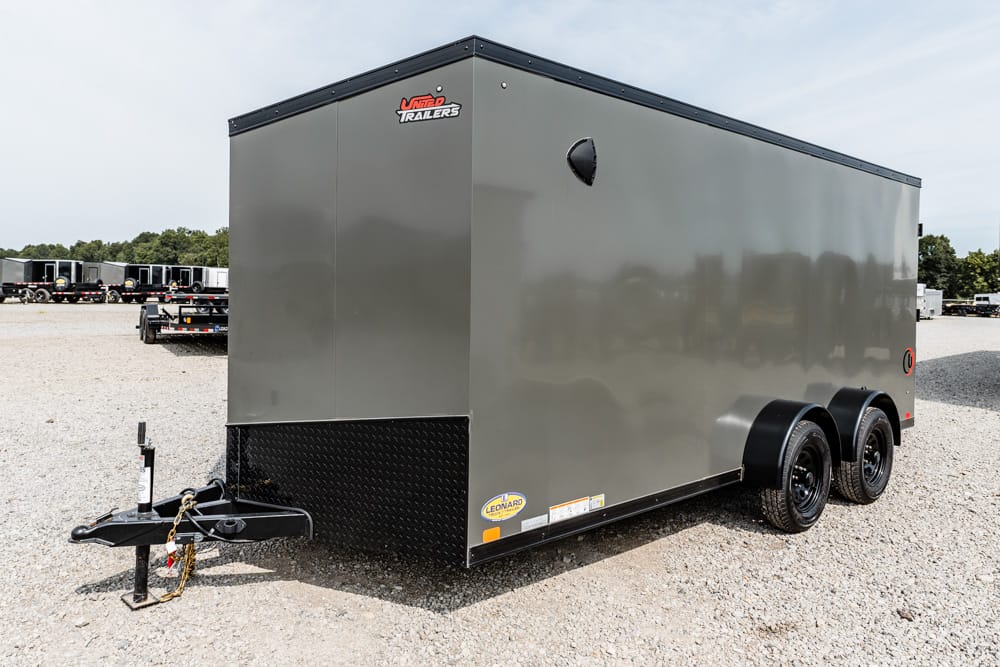 Grey enclosed dual-axle cargo trailer with a black diamond-plate front, parked on a gravel lot, hitch and safety chains visible at the front.