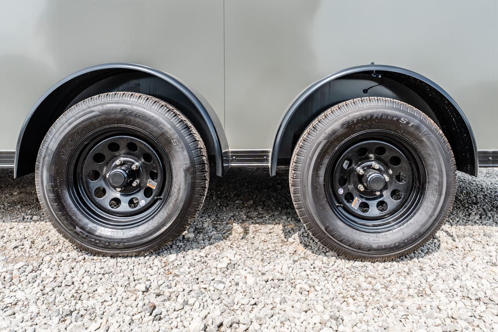 Two black steel trailer wheels with tires, under a gray trailer body on a gravel surface, showing wheel wells and hubs.