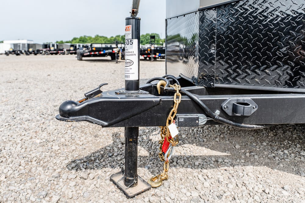 Close-up of a trailer hitch with its jack, safety chains, and padlock on a gravel lot, with trailers in the background.