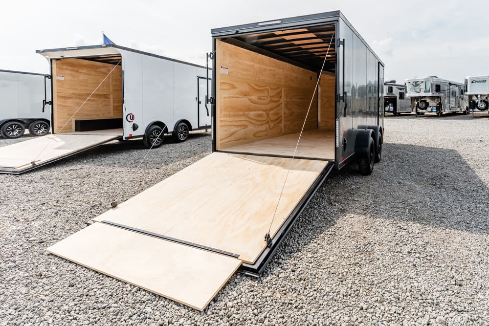Open cargo trailer with ramp down, showing plywood interior on a gravel lot with other trailers nearby.