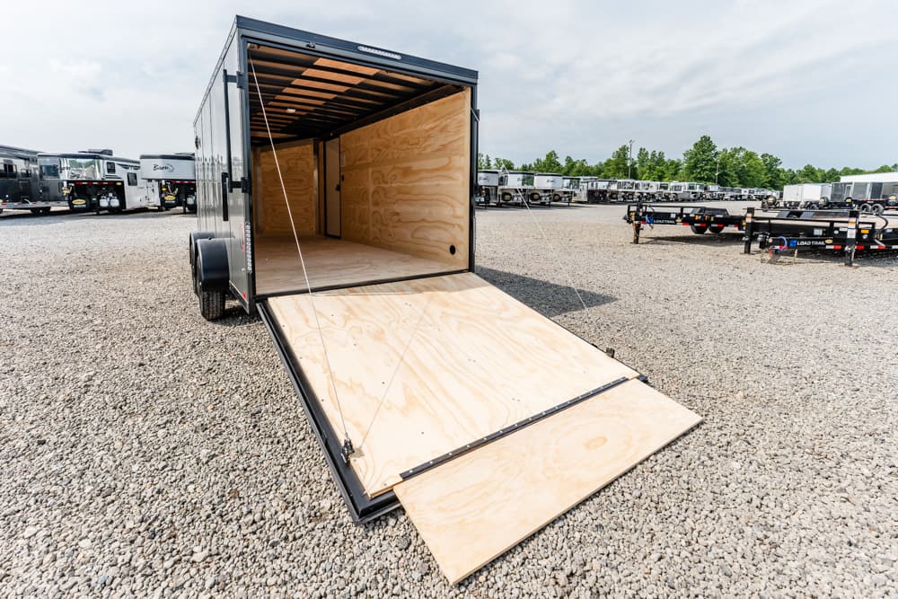Open black cargo trailer with a wooden ramp extended onto a gravel lot