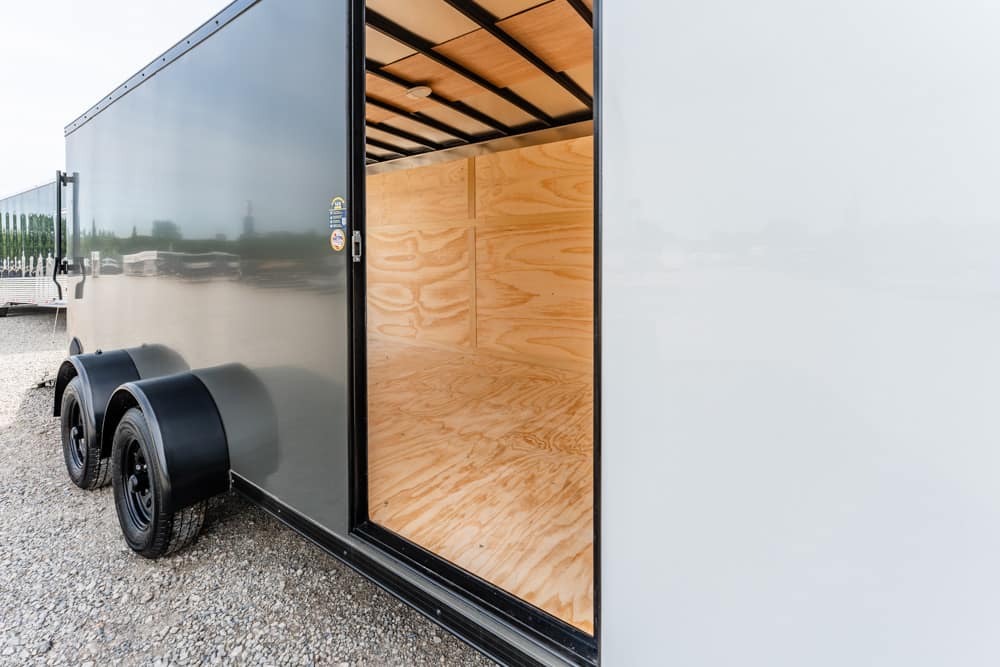 Open side door of a gray cargo trailer showing an unfinished plywood interior on a gravel surface.