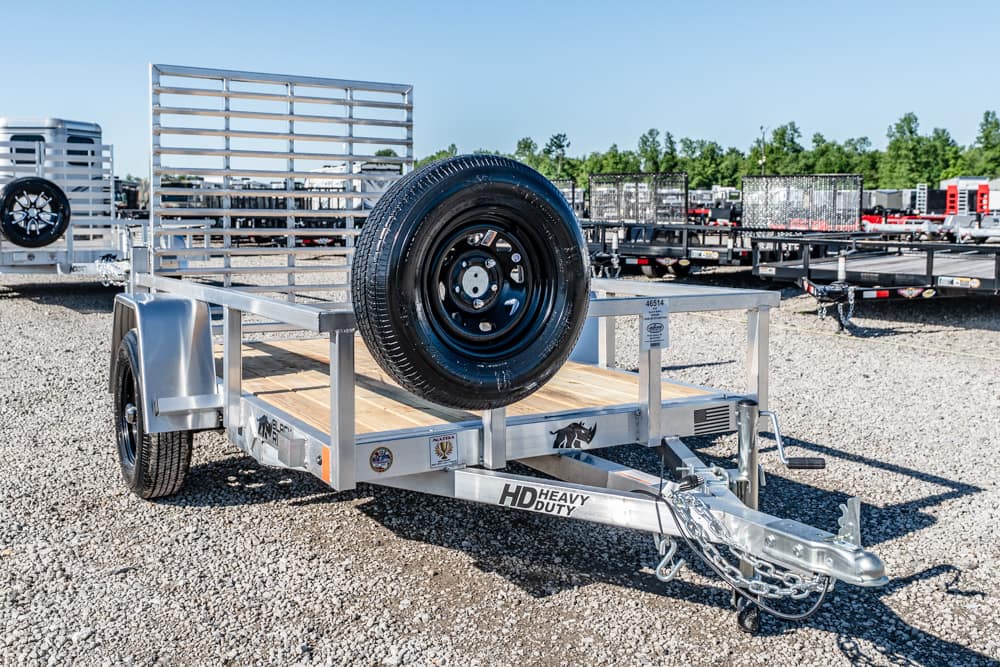 Silver HD Heavy Duty utility trailer with a wooden deck and mounted spare tire on a gravel lot, surrounded by other trailers and equipment.