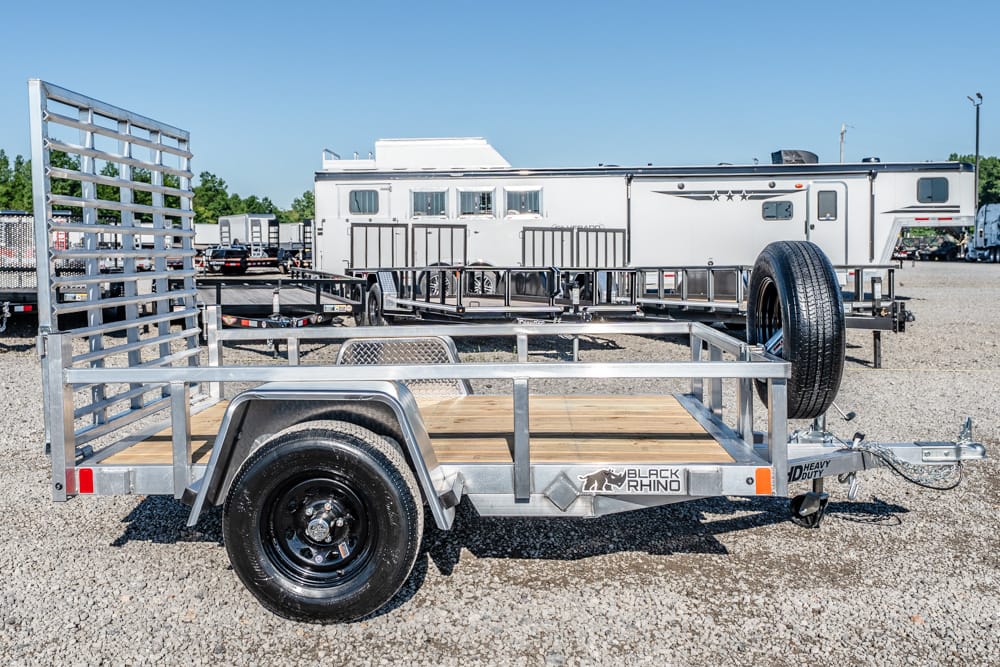 Galvanized utility trailer with wooden bed and spare tire, parked on a gravel lot with a large RV in the background.
