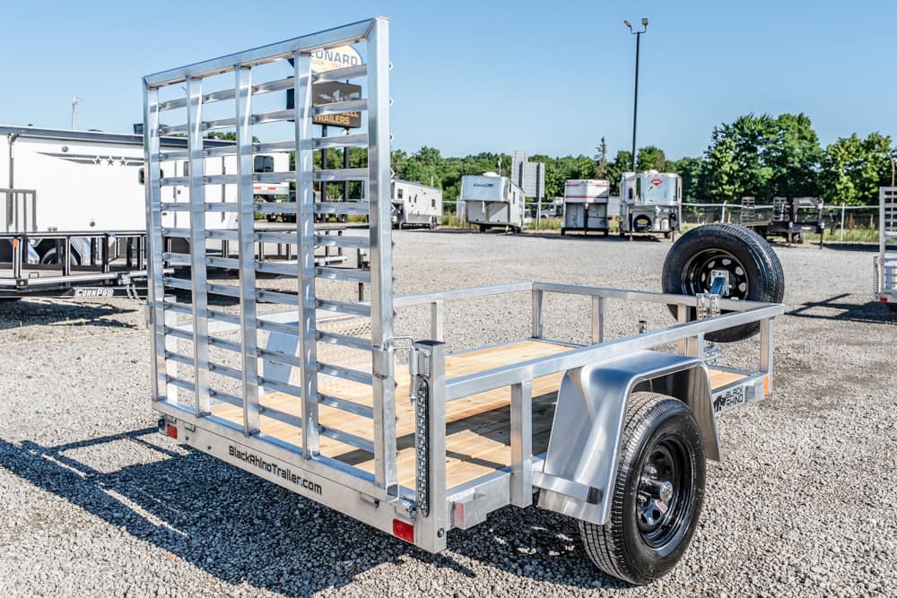 Metal utility trailer with a tall, slatted ramp gate, wooden deck, and spare tire in a gravel trailer yard