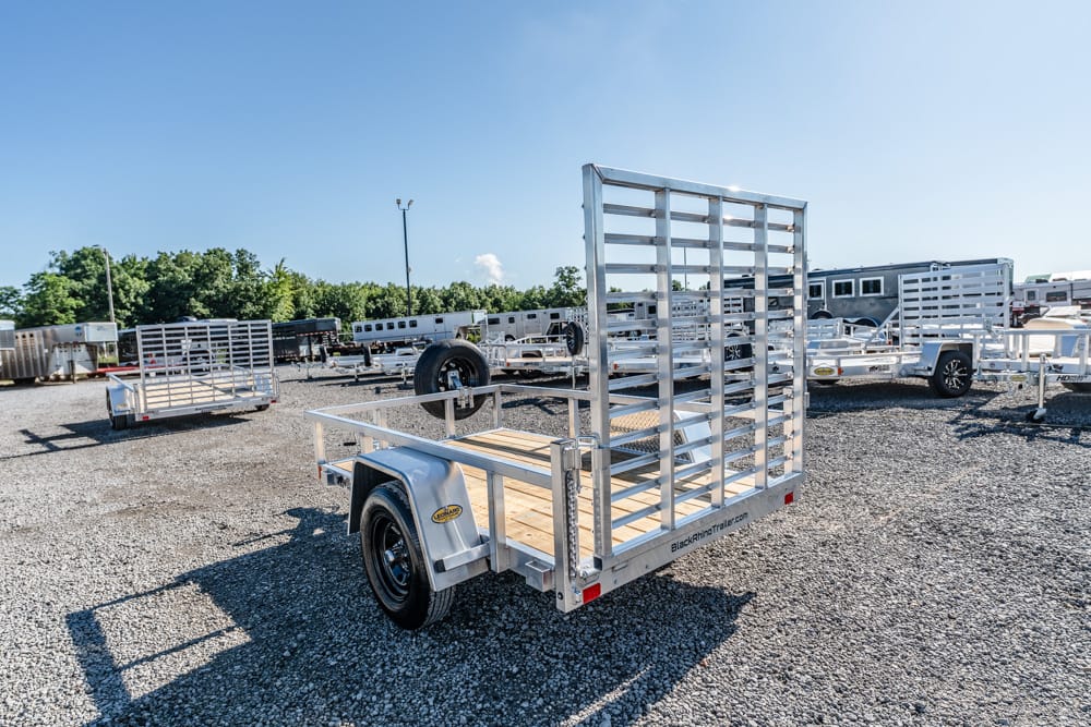 Aluminum utility trailer with a tall grated ramp gate on a gravel lot, surrounded by other trailers.